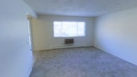 This single-floor space presents a straightforward, functional layout designed for comfortable everyday living. The main living area is finished with wall-to-wall carpeting in a neutral tone, contributing to a clean and understated aesthetic throughout. The room is illuminated by a large, horizontally oriented window that draws in natural light and offers a view of the surrounding neighborhood rooftops, lending the interior a bright and open feel despite its modest scale.

Climate control is well-considered, with both a wall-mounted air conditioning unit positioned beneath the window and a baseboard heating system running along the lower wall, ensuring year-round comfort. The walls are finished in a light, neutral color that enhances the brightness of the space and provides a versatile backdrop for various furnishing styles.

The room features a straightforward, open floor plan with minimal architectural detailing, making it practical and easy to arrange. The smooth ceiling and clean wall lines contribute to a simple, no-frills interior. An adjacent doorway suggests additional rooms or access points within the unit, indicating a functional multi-room layout suited to everyday residential use.