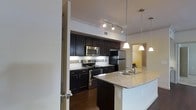 This contemporary kitchen showcases a sleek and functional design. Dark espresso-colored cabinetry provides a striking contrast against light walls and ceiling, creating a sophisticated ambiance. Stainless steel appliances, including a refrigerator, microwave, and oven with cooktop, offer both style and efficiency. The kitchen boasts a generous granite-topped island with a built-in sink, perfect for food preparation or casual dining. Overhead, a combination of recessed track lighting and elegant pendant lights illuminate the space effectively. A tiled backsplash adds texture and easy-to-clean practicality. The open layout extends to an adjacent room, hinting at a larger living area beyond. Hardwood flooring grounds the space, adding warmth to the overall modern aesthetic. This kitchen design seamlessly blends functionality with contemporary style, making it an inviting heart of the home.