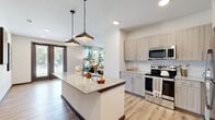 This single-floor residence is designed with a contemporary open-concept layout that promotes a seamless flow between the kitchen, dining, and living areas. The kitchen serves as a central hub, anchored by a granite-topped island with pendant lighting suspended on chain fixtures above. Light wood-tone cabinetry lines the walls alongside a subway tile backsplash, while stainless steel appliances—including a range, microwave, and refrigerator—complete the space. Light-toned hardwood-style flooring extends throughout the main living areas, complementing the neutral color palette.

The living room receives generous natural light through multiple windows dressed with sheer curtains, and a set of French doors provides direct access to an outdoor area. A ceiling fan adds to the room's comfort. Dark-trimmed interior doors add a contrasting design detail visible throughout the hallway connecting the main living spaces to the private rooms.

Two bedrooms each feature carpeted flooring, natural window light, and walk-in closets. Two bathrooms serve the residence—one with a glass-enclosed walk-in shower featuring marble-style tile surround, and the other with a soaking tub/shower combination. Both bathrooms include granite vanity countertops and light wood-tone cabinetry consistent with the overall design aesthetic.