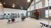Located at 380 Crown Dr in Emerson, GA, this well-appointed fitness center offers an impressive and fully equipped exercise environment across a single, expansive floor. The space is defined by its dramatic vaulted ceilings finished in a bold dark tone, which contrast elegantly with the white walls and light neutral carpeting below. Large multi-pane windows line the walls at multiple heights, flooding the interior with natural light while providing open views of the surrounding Georgia landscape.

The exercise room is thoughtfully organized to accommodate a wide range of fitness activities. The cardio zone features spin bikes with personal display screens, elliptical trainers, treadmills, and stair climbers positioned near the windows to take full advantage of the outdoor views. The strength training area is equally well-equipped, offering selectorized weight machines, cable systems, a Smith machine with an adjustable bench, a full dumbbell rack, and a functional training station stocked with kettlebells, medicine balls, and resistance bands. A floor-to-ceiling mirror anchors one section of the room, enhancing the sense of space and supporting form-focused training.

Warm pendant lighting complements the natural light, while elegant wood-paneled accents and double glass entry doors add a refined, upscale character to this community fitness facility.