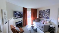 This single-floor space offers a warm and inviting atmosphere, thoughtfully designed with a blend of comfort and contemporary style. The living area serves as the focal point of the space, anchored by a tiled fireplace that adds both character and coziness to the room. A wall-mounted flat-screen television is positioned above the fireplace, creating a practical and visually balanced entertainment setup.

Natural light pours into the space through a sliding glass door framed by striking burnt-orange floor-to-ceiling curtains, which add a bold pop of color and a sense of height to the room. The door opens to what appears to be an outdoor balcony or patio area, offering a connection to the exterior.

The living area is furnished with a comfortable grey sofa adorned with patterned accent pillows, complemented by a sleek glass-top coffee table with a sculptural base that sits atop a plush area rug. A leather accent chair rounds out the seating arrangement, adding texture and warmth to the space.

The walls feature tasteful artwork, including a large mixed-media canvas that adds visual interest and personality. The neutral wall tones and light ceiling allow the warm furnishings and decor accents to take center stage, resulting in a cohesive and welcoming living environment.