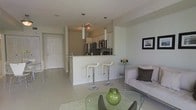This well-appointed single-floor space showcases a clean, modern design with an open-concept layout that seamlessly connects the kitchen, dining, and living areas. The kitchen is outfitted with stainless steel appliances, white cabinetry, and a light-toned countertop that extends into a breakfast bar, complete with two contemporary white bar stools — perfect for casual dining or entertaining. Pendant lighting with sleek cylindrical shades hangs above the bar, adding a refined touch to the kitchen area. Track lighting further illuminates the space with a warm, functional glow. The living area features a light grey sofa accented with dark decorative pillows, a glass-top coffee table, and a small side table holding stylish glassware. A framed piece of wall art adds a subtle decorative element. Light-toned tile flooring runs throughout, enhancing the bright and airy atmosphere. A white entry door is visible down the hallway, contributing to the open, flowing feel of the floor plan. Overall, the space presents a cohesive, contemporary aesthetic with thoughtful design details and a neutral color palette that feels both inviting and functional.