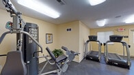 Located at 31275 Haggerty Rd in Novi, MI, this single-floor fitness center is designed to provide a comprehensive exercise environment across several interconnected rooms. The facility features warm yellow-toned walls and dark rubber and carpet flooring throughout, creating a clean, functional atmosphere well-suited for physical activity.

The main workout area is generously sized, housing multiple cardio machines including treadmills, elliptical trainers, and a stationary recumbent bike. A multi-station cable weight machine and an abdominal/leg press apparatus provide strength training options, complemented by instructional exercise posters mounted on the walls. Flush-mounted fluorescent lighting ensures the space is consistently and evenly lit.

A dedicated cardio room accommodates two side-by-side treadmills positioned near a window with natural light filtering through slatted blinds. An additional room features a stationary upright bike, a wall-mounted flat-screen television, and a corner plant, creating a more relaxed setting for low-intensity workouts. A water fountain and waste receptacle are conveniently accessible near the corridor connecting the rooms.

The facility includes clearly marked exits, ventilation systems, and access to adjoining hallways, contributing to an organized and safe workout environment overall.
