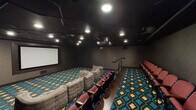 This single-level private home theater is designed to deliver an immersive cinematic experience. The room is anchored by a large projection screen mounted on the front wall, paired with a ceiling-mounted projector positioned toward the rear of the space. The dark, acoustically treated walls and matte black ceiling work together to minimize light reflection and enhance viewing conditions, while recessed can lighting provides ambient illumination when needed.

Seating is divided into two distinct zones. The lower level features a generous arrangement of modular, tufted sofas in a neutral gray upholstery, configured in a curved layout for relaxed, lounge-style viewing. Elevated on a raised platform along the sides and rear of the room, traditional theater-style seats upholstered in deep red offer a more formal, stadium-like viewing experience, with a wooden step separating the two levels.

Throughout the room, a bold teal, brown, and gold geometric patterned carpet adds a vibrant visual contrast to the otherwise dark interior. Wall paneling displays a subtle herringbone texture, contributing to the refined yet dramatic atmosphere of the space. A central aisle and handrail facilitate easy navigation between seating sections.