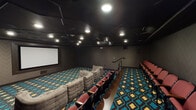 This single-level private home theater is designed to deliver an immersive cinematic experience. The room is anchored by a large projection screen mounted on the front wall, paired with a ceiling-mounted projector positioned toward the rear of the space. The dark, acoustically treated walls and matte black ceiling work together to minimize light reflection and enhance viewing conditions, while recessed can lighting provides ambient illumination when needed.

Seating is divided into two distinct zones. The lower level features a generous arrangement of modular, tufted sofas in a neutral gray upholstery, configured in a curved layout for relaxed, lounge-style viewing. Elevated on a raised platform along the sides and rear of the room, traditional theater-style seats upholstered in deep red offer a more formal, stadium-like viewing experience, with a wooden step separating the two levels.

Throughout the room, a bold teal, brown, and gold geometric patterned carpet adds a vibrant visual contrast to the otherwise dark interior. Wall paneling displays a subtle herringbone texture, contributing to the refined yet dramatic atmosphere of the space. A central aisle and handrail facilitate easy navigation between seating sections.