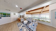 Located at 300 W Airport Blvd in Sanford, Florida, this single-floor communal space offers two thoughtfully designed living areas that cater to both social gatherings and quiet leisure. The first living area is a bright, open lounge characterized by soaring vaulted ceilings with exposed wood beams and tongue-and-groove wood paneling, flooding the space with warmth and architectural charm. Large windows line the walls, offering lush outdoor views and abundant natural light. The room features durable wood-look flooring, a decorative chandelier, and a seating arrangement of floral-patterned armchairs and sofas centered around a white coffee table atop a coastal-inspired area rug. A built-in bookshelf and a small reading nook add functional charm, while colorful artwork and vibrant potted plants complete the inviting atmosphere. A stainless steel elevator provides convenient access throughout. The second living area serves as a dedicated media lounge with a more intimate, enclosed feel. Neutral-toned recliner chairs are arranged around a large wall-mounted television, set against a darker accent wall. Patterned carpeting, recessed lighting, and a popcorn machine accent this comfortable screening space. Together, these two rooms create a versatile, well-appointed environment suited for community living.