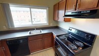 This well-appointed two-bedroom, one-bathroom residence offers a functional and comfortable single-floor layout. The upgraded kitchen is a standout feature, showcasing rich cherry cabinetry paired with sleek dark countertops and a full suite of black appliances, including a coil-top range, dishwasher, refrigerator, and over-range exhaust hood. A window above the sink fills the kitchen with natural light. A cherry-finished peninsula with dark countertop separates the kitchen from the carpeted living area, creating an open and connected feel. The living space is bright and open, with a large window and wall-unit air conditioning. Both bedrooms are carpeted and feature windows with outdoor views, baseboard heating, and ample closet storage, including a built-in closet with shelving. One bedroom is enhanced by a ceiling fan with light fixture. The bathroom is clean and bright, finished with white cabinetry, a large vanity mirror, wall sconce lighting, and vinyl plank flooring. Light-toned walls and neutral carpet throughout contribute to a cohesive, inviting ambiance across the entire residence.