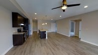 This single-floor space showcases a thoughtfully designed open-concept layout that seamlessly blends the kitchen and living areas into one inviting, cohesive environment. The kitchen is a standout feature, equipped with dark cabinetry, granite countertops, stainless steel appliances, and a central island with a built-in sink, illuminated by elegant pendant lights that hang overhead. Recessed lighting throughout the space ensures consistent, well-distributed brightness across every corner. The warm, wood-style luxury vinyl plank flooring extends throughout the main living area, lending a clean and contemporary feel underfoot. Neutral-toned walls complement the overall palette, creating a bright and welcoming ambiance. A ceiling fan with an integrated light fixture is centrally positioned in the living area, adding both functional ventilation and a finishing decorative touch. The open floor plan allows for generous, flexible use of the living space, while doorways visible in the background hint at additional rooms beyond, such as bedrooms or utility spaces. White trim and baseboards provide crisp contrast against the neutral walls, adding a polished, finished look throughout. The overall design balances modern aesthetics with everyday practicality.