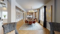 This thoughtfully designed community room spans a single floor and offers a variety of gathering spaces suited for both relaxed socializing and active entertainment. The main lounge area makes an immediate impression with its soaring coffered ceiling, an ornate amber pendant chandelier, and a stone-accented fireplace flanked by a wall-mounted television — creating a warm, inviting focal point. Plush seating and herringbone-patterned hardwood flooring carry throughout the space, providing a cohesive and refined aesthetic. Large windows dressed with sheer and blackout drapery allow ample natural light while framing outdoor views. Adjacent to the lounge, a formal dining area features a round dark-top table surrounded by leather chairs beneath a brass chandelier, set atop a layered area rug. A dedicated game nook is highlighted by striking geometric patterned wallcovering, a wall-mounted oversized Scrabble board, a chalkboard, curated whimsical framed artwork, and cozy lounge seating with a starburst ceiling fixture. The kitchen area is outfitted with contemporary flat-front cabinetry in a warm taupe finish, sleek quartz countertops, full subway tile backsplash extending to the ceiling, stainless steel appliances, and stylish smoked glass pendant lighting — making it fully functional for community events and gatherings.