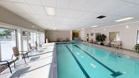 This indoor aquatic facility is thoughtfully designed to accommodate both recreational swimmers and serious lap enthusiasts. The centerpiece is a generously sized multi-lane lap pool with clearly marked lanes, finished in calming aqua-toned tiles that reflect the overhead lighting beautifully. Adjacent to the lap pool is a raised hot tub, accessible via steps with handrail support. The facility is enclosed within a clean, single-floor structure featuring a suspended tile ceiling with recessed and panel lighting that ensures consistent illumination throughout.

Along the poolside, seating areas with lightweight chairs are arranged for observers or resting swimmers, and lush potted trees add warmth and a touch of greenery to the otherwise neutral, light-toned walls. The surrounding deck is finished with patterned tile flooring that provides both aesthetic appeal and practical slip resistance.

Large windows and glass doors along one side of the facility allow natural light to pour in and offer views of an outdoor pool area, which is accessible directly through the building. Additional utility spaces, including what appears to be a changing or equipment room, are accessible via interior doors. Signage with pool rules and safety information is posted prominently, underscoring the facility's commitment to a safe and organized environment.