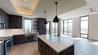 This single-floor high-rise residence showcases a thoughtfully designed open-concept layout with modern finishes and sweeping urban views. The expansive living area is defined by floor-to-ceiling glass windows on multiple walls, allowing abundant natural light to pour in and offering panoramic cityscape views. Exposed concrete columns add an industrial architectural element, while light hardwood flooring and a ceiling fan contribute to the space's airy, contemporary feel.

The chef's kitchen is a standout feature, outfitted with dark wood cabinetry, a large center island with a white quartz countertop, and stainless steel appliances including a refrigerator, range, microwave, and dishwasher. A stainless steel farmhouse sink sits beneath a window framing additional city views. Geometric black pendant lights with gold interiors hang above the island, complementing the kitchen's tray ceiling detail.

The bedrooms are carpeted and well-lit, with large windows offering elevated urban views and ceiling fans for comfort. The primary bathroom features a glass-enclosed shower with decorative geometric tile, a dual-sink vanity with dark cabinetry, pendant lighting, and light tile flooring. A secondary bathroom includes a soaking tub with tiled surround, a single-sink vanity, and recessed lighting, maintaining the same refined aesthetic throughout.