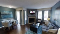 This single-floor space showcases an open-concept layout that seamlessly connects the living and dining areas, creating a cohesive and functional environment. The living area is anchored by a corner fireplace with a light tile surround and white mantel, above which a wall-mounted flat-screen television is positioned for optimal viewing. The accent wall behind the fireplace is finished in a deep charcoal gray, providing a bold contrast to the lighter surrounding walls and adding visual depth to the room.

The flooring throughout is rich, dark hardwood, lending warmth and continuity across both the living and dining zones. A teal geometric-patterned area rug defines the seating area, complementing the neutral gray upholstered furniture. Multiple windows dressed with gray grommet-top curtains allow generous natural light to filter into the space, with a sliding glass door visible on one side offering additional outdoor access.

The dining area, positioned adjacent to the living room, features a round table with modern chairs, making efficient use of the open floor plan. A textured popcorn ceiling completes the interior, while the overall color palette of grays, teals, and warm wood tones creates a balanced, contemporary ambiance throughout.