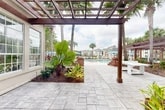 Located at 9800 Touchton Rd in Jacksonville, Florida, this generously sized outdoor amenity facility delivers a resort-style experience within a lush, tropical setting. The centerpiece is a large, sparkling swimming pool framed by tall palm trees and surrounded by an expansive pool deck outfitted with lounge chairs and shade umbrellas, ideal for relaxing in the Florida sunshine.

A striking brick-accented clubhouse anchors the space, its classic architectural design complemented by dormer windows, shuttered facades, and a covered pergola structure with a dark wood lattice roof. The pergola-shaded patio features elegant stone tile flooring and serves as a comfortable transition between the building and the pool area, with lush tropical plantings — including large elephant ear plants and colorful foliage — adding vibrant greenery throughout.

A charming wooden boardwalk with white railings and rich wood-tone decking winds through manicured landscaping, providing scenic views of a neighboring pond and the broader community. The sun deck area offers additional lounge seating beneath blue market umbrellas, bordered by carefully maintained shrubs and palm trees. Thoughtfully designed pathways connect each zone of this outdoor retreat, creating a cohesive and inviting environment that emphasizes natural beauty, open-air comfort, and a distinctly Floridian ambiance.