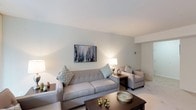 This single-floor residence offers a thoughtfully arranged layout with a cohesive, neutral aesthetic throughout. The living room serves as the central gathering space, featuring plush wall-to-wall carpeting, soft white walls, and large sliding glass doors dressed with vertical blinds that allow generous natural light to filter in. The room comfortably accommodates a full seating arrangement, including a tufted sofa, accent chairs, and a media console.

Adjacent to the living area, the dining room provides an intimate setting highlighted by a ceiling fan with a light fixture overhead and a vertical-blind-covered window that brings in additional daylight. The kitchen is functional and well-equipped, featuring warm wood cabinetry, dark granite-toned countertops, white appliances including a gas range and refrigerator, and light tile flooring. A compact dining nook within the kitchen offers a casual eating option.

Both bedrooms are carpeted and serene, each featuring vertical blinds, ample wall space, and a calm, restful atmosphere. The primary bedroom includes space for a dresser and a sitting chair. The bathroom is clean and practical, with a white vanity, wood cabinetry, a well-lit mirror above the sink, and a shower-tub combination.