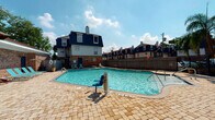 This outdoor amenity area in Gretna, Louisiana, is designed to serve as a central recreational hub for its surrounding residential community. The centerpiece is a generously sized swimming pool with clear, blue water, surrounded by a paved brick patio that provides ample space for lounging. Depth markers are clearly visible along the pool's edge, and an ADA-compliant pool lift ensures accessibility for all residents. Pool-side seating is available along the perimeter, complemented by a covered pergola structure that offers a shaded retreat. The pergola features warm wood beam construction with a corrugated roof panel and is furnished with comfortable wicker sectional seating, creating an inviting outdoor living area directly adjacent to the pool. The patio surface throughout is composed of interlocking pavers in a neutral tone, lending a cohesive and polished appearance to the space. A black wrought-iron security fence encloses the area, with a gated entry providing controlled access. Just beyond the fence, a playground with colorful equipment is visible, adding family-friendly amenities to the community. Palm trees and landscaping accent the surroundings, contributing to the relaxed, subtropical atmosphere characteristic of the greater New Orleans area.
