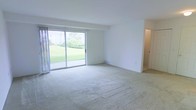 This single-floor space offers a practical and comfortable layout designed to maximize everyday living. The kitchen is efficiently arranged with wood-trimmed white cabinetry spanning both upper and lower sections, providing generous storage for all culinary essentials. It is equipped with an electric coil range, an overhead ventilation hood, and a full-size refrigerator. A stainless steel sink is positioned at the counter's edge, and a doorway from the kitchen leads conveniently to additional areas of the unit.

The living area is a standout feature of the space, characterized by its open, airy feel and neutral wall-to-wall carpeting that adds warmth underfoot. Large sliding glass doors dominate one wall, flooding the room with abundant natural light and offering an unobstructed view of a well-maintained green lawn with lush landscaping. The sliding doors also provide direct access to the outdoor area, seamlessly blending interior and exterior living. The clean white walls and simple architectural lines throughout the space create a bright, neutral canvas well-suited for a variety of décor styles. Overall, the layout is straightforward and functional, making efficient use of its single-floor footprint.