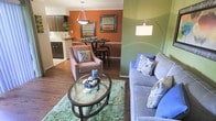 This single-floor residence offers a thoughtfully designed open-concept layout that seamlessly connects the living, dining, and kitchen areas into one cohesive and inviting space. The living area is anchored by rich hardwood-style flooring and features a sage green accent wall that adds a fresh, energetic tone to the room. A plush area rug, glass-top oval coffee table, and a comfortable upholstered sofa create a relaxed yet stylish seating arrangement, enhanced by a modern arc floor lamp that provides warm ambient lighting. Adjacent to the living space, the dining area is defined by a striking burnt orange accent wall that lends warmth and character to the space. A round bistro-style dining table with bar stools sits beneath a classic pendant light fixture, while framed artwork on the wall adds a decorative finishing touch. The kitchen, visible through the open pass-through, features dark cabinetry, light countertops, and functional layout. The open flow between all three areas makes the space feel expansive and well-connected, ideal for both everyday living and entertaining. The bold use of color throughout, paired with coordinated furnishings and thoughtful décor, gives this residence a lively and well-curated aesthetic.