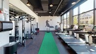 This contemporary fitness center is thoughtfully designed to accommodate a wide range of workout styles within a single, open-plan floor. The space features an exposed industrial ceiling with dark-painted ductwork, pendant lighting, and structural concrete columns that contribute to a sleek, modern aesthetic.

The main workout floor is finished with durable rubber flooring and is organized into distinct training zones. A row of treadmills lines the windowed wall, while elliptical trainers and a spin bike occupy a dedicated cardio area bathed in natural light from large, floor-to-ceiling windows that offer views of the surrounding neighborhood. A green turf lane runs through the center of the space, ideal for functional and sled-based training.

Strength training is well-supported with a multi-station cable machine, a weight bench, a full rack of dumbbells, and additional free weights and barbells. A separate studio area features deep burgundy accent walls, full-length mirrors with a ballet barre, medicine balls, and a mounted display screen — suitable for barre, stretching, or guided training sessions. A branded fox logo mural adorns one wall, adding a distinctive visual identity to the space. Ample natural light and thoughtful equipment placement make this fitness center both functional and visually engaging.