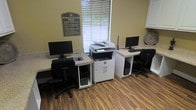 This single-floor business center in Dallas, TX, is a functional and efficiently organized workspace designed to meet essential office needs. The room features two dedicated computer workstations, each equipped with a desktop PC, monitor, keyboard, and ergonomic rolling chair, allowing multiple users to work simultaneously. Positioned centrally between the workstations is a large multifunction printer and copier, providing convenient access to printing, scanning, and copying services from either station.

The workstation surfaces feature a speckled granite-style laminate finish, offering a clean and durable work area. A posted "Business Center Rules" sign on the sage-green wall ensures orderly use of the shared space. Natural light filters into the room through a sizable window fitted with horizontal blinds, creating a bright yet controlled environment. Wood-look plank flooring adds warmth to the utilitarian setting, while white cabinetry along the upper wall provides additional storage. The overall layout is straightforward and practical, making it a reliable resource for those needing access to office equipment and computing facilities.