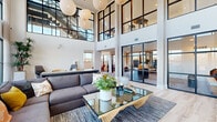 This contemporary multi-story amenity space is defined by its dramatic double-height ceilings and expansive floor-to-ceiling windows framed in sleek black steel, flooding the interiors with natural light and offering sweeping views of the surrounding area. Light wood flooring runs throughout, complementing the crisp white walls and modern industrial aesthetic.

The main social lounge serves as the centerpiece, featuring a generous seating area with plush sofas, a sculptural glass coffee table, and a cluster of oversized woven globe pendant lights that add warmth and visual interest. An open mezzanine level, accessible via a striking steel staircase with cable railings, overlooks the lounge below and connects the floors with an airy sense of openness.

A dedicated game room provides entertainment with a shuffleboard table and ping-pong table set against light hardwood flooring and large windows with outdoor views. The coworking and lounge area offers a thoughtful mix of private seating nooks framed in warm wood structures, casual lounge seating, and a glass-enclosed conference room equipped with ergonomic seating—ideal for focused work or collaborative meetings.

Throughout the space, carefully curated furnishings, accent walls in deep navy, and pops of terracotta and mustard create an inviting, design-forward atmosphere that balances functionality with comfort across all three floors.