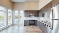 This well-appointed single-floor unit showcases a thoughtful modern design with consistent finishes throughout. The kitchen serves as a standout centerpiece, featuring floor-to-ceiling dark wood-grain cabinetry, granite countertops, a white tile backsplash, and a full suite of stainless steel appliances including a gas range, dishwasher, and refrigerator. Soaring ceilings in the kitchen and living areas create an open, airy atmosphere, while large windows invite abundant natural light and outdoor views. Wood-look plank flooring runs seamlessly throughout the entire unit, lending warmth and visual continuity to every room. The hallway connects multiple well-proportioned bedrooms, each featuring tall ceilings, large windows with exterior views, and ample closet space. At least one bedroom benefits from a private en-suite bathroom. The bathrooms are finished with granite-topped vanities, dark wood cabinetry with brushed nickel hardware, and clean white walls for a polished look. The overall palette of warm beige and neutral grey tones, paired with crisp white trim and panel doors, creates a cohesive, contemporary aesthetic throughout the space. The layout is practical and well-organized, offering a comfortable and modern living environment.