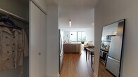 This contemporary single-floor residence offers a thoughtfully designed open-concept layout that seamlessly connects the kitchen, dining, and living areas. The kitchen is a standout feature, equipped with stainless steel appliances including a refrigerator, electric range, microwave, and dishwasher, complemented by a large central island with a white quartz countertop, a herringbone tile backsplash, and a combination of white upper cabinets and warm wood-toned lower cabinets. Rich hardwood flooring flows throughout the entire unit, lending warmth and continuity to each space. The living area is bathed in natural light from floor-to-ceiling windows that frame sweeping panoramic views of the city skyline and waterfront beyond. The bedroom continues this connection to the outdoors with its own expansive window wall offering impressive elevated city views, and includes a built-in closet for ample storage. The bathroom is finished with a floating wood-grain vanity, white quartz countertop, a mirrored medicine cabinet, hexagonal tile flooring, and a shower/tub combination. An in-unit laundry area provides added convenience. The overall ambiance is clean, bright, and modern, with a neutral palette that highlights the architectural details and the stunning views.