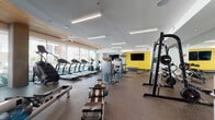 Located at 6001 S Congress Ave in Austin, TX, this modern fitness center is designed to support a comprehensive workout experience. The space is anchored by a wall of floor-to-ceiling windows and glass doors that flood the interior with natural light and offer views of the surrounding outdoor area, creating an open and energizing atmosphere. A warm wood-paneled ceiling detail adds an inviting architectural contrast to the otherwise clean, contemporary white walls and durable rubber flooring throughout.

The cardio zone is well-stocked with treadmills, a StairMaster, ellipticals, and WaterRower rowing machines, all positioned to take advantage of the natural light. Strength training is thoroughly covered with cable machines, weight racks, and adjustable benches spread across the main floor. A dedicated TRX Training Zone features a full functional training rig complete with suspension trainers, medicine balls, and resistance equipment, set against a bold, colorful graphic wall mural that adds visual energy to the space.

A separate area provides a more relaxed setting with wall-mounted flat-screen TVs, built-in wooden shelving, and a yellow accent wall that injects a vibrant pop of color. Ceiling fans and linear LED lighting ensure comfort and visibility throughout. The fitness center offers a thorough, amenity-rich environment suited for all fitness levels.