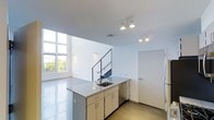 This contemporary two-story space showcases a clean, minimalist design characterized by crisp white walls, polished concrete floors, and an abundance of natural light. The open-plan main level centers around a kitchen island finished with a granite-style countertop, an undermount sink, and warm-toned cabinetry beneath. Track lighting overhead provides focused illumination throughout the space. A striking staircase with a sleek black metal railing connects the two floors, serving as both a functional and architectural feature. Large, multi-pane windows along one wall invite generous amounts of natural light, offering views of surrounding greenery and reinforcing the bright, airy ambiance. Wall-mounted accent lighting adds a subtle layer of warmth alongside the recessed fixtures above. The overall aesthetic is decidedly modern and uncluttered, with an emphasis on clean lines and open volume. The second floor is accessible via the contemporary staircase, expanding the living or working potential of the space. The combination of industrial-inspired finishes — polished concrete underfoot, exposed ceiling details, and minimal ornamentation — creates a versatile environment suited to a variety of uses.