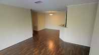 This single-floor residence offers a clean, open layout designed for comfortable everyday living. The main living area is a standout feature, showcasing rich, dark hardwood-style flooring that runs throughout and adds warmth and visual continuity to the space. Neutral, light-toned walls complement the flooring and create a bright, welcoming atmosphere throughout.

A defining architectural element is the curved pass-through counter that connects the living area to the adjacent kitchen, enhancing the open feel while maintaining a subtle separation between spaces. This countertop detail features clean curved edges and is supported by a structural column, adding a functional yet stylish design touch.

The textured popcorn ceiling adds classic character overhead, while a centrally hung pendant light fixture provides focused illumination to the dining or transitional area. Visible HVAC vents in both the ceiling and lower wall areas ensure efficient climate control throughout the space.

Crown molding accents are visible near the pass-through area, lending a refined finishing detail to the interior. The open flow between rooms makes the space feel expansive and versatile, lending itself well to a variety of furniture arrangements. Overall, the space presents a practical and inviting environment with consistent finishes and a cohesive, neutral design palette.