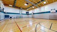 Located at 25 Woodridge Crescent in Ottawa, this expansive gymnasium is part of the Accora Centre community facility. The single-floor space offers a generously sized, multi-use hardwood sports court finished to a high standard, with clearly marked lines for basketball and other court sports. The Accora Centre logo is prominently displayed at center court, reinforcing the facility's identity.

The gymnasium features soaring ceilings supported by exposed wood truss construction, lending the space a warm, structural aesthetic while providing ample vertical clearance for athletic activities. Multiple wall-mounted and retractable basketball hoops are positioned along both ends and sides of the court, making the space adaptable for a variety of game configurations.

The walls are finished in white concrete block with a distinctive teal tile accent band running at mid-height, creating a clean and functional visual boundary. High, clerestory-style windows line the upper walls, allowing natural light to filter in while maintaining focus and minimizing glare on the court surface. Bright overhead fluorescent lighting ensures consistent illumination throughout.

Padded wall mats are mounted along key perimeter sections for player safety, and double entry doors with exit signage provide clear access points. Security cameras and a fire suppression system are also installed, reflecting the facility's commitment to safety and operational readiness.