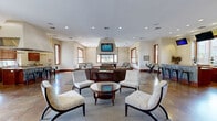 Located at 1200 Harrison Creek Blvd in Petersburg, Virginia, this generously sized single-story community clubhouse offers a well-appointed gathering space designed for comfort and socializing. The open-concept layout is anchored by polished, stained concrete floors that lend the space a warm, contemporary character throughout.

The main lounge area is furnished with a large sectional sofa, accent chairs, and a central coffee table arranged on a neutral area rug, creating an inviting setting for relaxation. Recessed lighting and multiple wood-trimmed windows allow ample natural light to fill the room. A wall-mounted television serves as a focal point for the seating area.

Adjacent to the lounge is a dedicated recreation zone featuring a full-size pool table and additional seating, ideal for entertainment and leisure. The space flows seamlessly into a fully equipped kitchen boasting rich mahogany cabinetry, granite countertops, a large center island with bar seating, stainless steel appliances including a double wall oven and gas cooktop, and a dramatic overhead range hood that rises toward the soaring ceiling. A dedicated café station with cabinetry, granite counters, and a coffee setup provides a convenient refreshment area. The combination of thoughtful amenities and open, connected spaces makes this clubhouse an exceptional community hub.