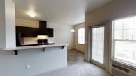 This single-floor residence offers a thoughtfully designed open-concept layout that seamlessly connects the kitchen, dining, and living areas. The kitchen is outfitted with rich, dark espresso cabinetry that provides generous storage space, complemented by a black gas range with an overhead microwave hood. A light-toned breakfast bar extends from the kitchen peninsula, offering bar-style seating and a natural transition into the adjoining living space, which features plush carpet flooring. A pendant light fixture hangs above the dining area, adding a warm focal point to the space. The dining area is finished with tile flooring and benefits from abundant natural light streaming through a large window and a full-length glass door fitted with blinds, which also provides convenient exterior access. The neutral wall tones and white ceilings throughout create a clean, bright ambiance that enhances the open feel of the layout. The combination of carpet in the living area and tile in the dining zone adds practical definition between the two spaces while maintaining a cohesive aesthetic. Overall, the space delivers a functional and welcoming environment well-suited for everyday living.