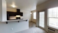 This single-floor residence offers a thoughtfully designed open-concept layout that seamlessly connects the kitchen, dining, and living areas. The kitchen is outfitted with rich, dark espresso cabinetry that provides generous storage space, complemented by a black gas range with an overhead microwave hood. A light-toned breakfast bar extends from the kitchen peninsula, offering bar-style seating and a natural transition into the adjoining living space, which features plush carpet flooring. A pendant light fixture hangs above the dining area, adding a warm focal point to the space. The dining area is finished with tile flooring and benefits from abundant natural light streaming through a large window and a full-length glass door fitted with blinds, which also provides convenient exterior access. The neutral wall tones and white ceilings throughout create a clean, bright ambiance that enhances the open feel of the layout. The combination of carpet in the living area and tile in the dining zone adds practical definition between the two spaces while maintaining a cohesive aesthetic. Overall, the space delivers a functional and welcoming environment well-suited for everyday living.