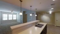 This single-floor residence is designed with an open-concept layout that promotes a natural flow between the kitchen and living areas. The kitchen serves as a central hub, anchored by an expansive light-colored countertop that doubles as a breakfast bar, offering casual seating and a clear sightline into the adjoining living space. A double stainless steel sink is neatly integrated into the countertop, accompanied by a classic chrome faucet. Three pendant lights hang in a row above the counter, providing focused task lighting while contributing a refined visual element to the space. Dark cabinetry beneath the counter adds a grounding contrast to the light surfaces above. A track lighting fixture on the ceiling extends illumination further into the room. The living area features plush carpeted flooring and is bathed in natural light from large windows dressed with horizontal blinds. Neutral-toned walls throughout create a calm, cohesive palette. Multiple doorways visible from the main living area hint at additional rooms, each framed cleanly with white paneled doors. The overall ambiance is clean, contemporary, and functional, offering a comfortable and well-organized single-level living environment.