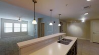 This single-floor residence is designed with an open-concept layout that promotes a natural flow between the kitchen and living areas. The kitchen serves as a central hub, anchored by an expansive light-colored countertop that doubles as a breakfast bar, offering casual seating and a clear sightline into the adjoining living space. A double stainless steel sink is neatly integrated into the countertop, accompanied by a classic chrome faucet. Three pendant lights hang in a row above the counter, providing focused task lighting while contributing a refined visual element to the space. Dark cabinetry beneath the counter adds a grounding contrast to the light surfaces above. A track lighting fixture on the ceiling extends illumination further into the room. The living area features plush carpeted flooring and is bathed in natural light from large windows dressed with horizontal blinds. Neutral-toned walls throughout create a calm, cohesive palette. Multiple doorways visible from the main living area hint at additional rooms, each framed cleanly with white paneled doors. The overall ambiance is clean, contemporary, and functional, offering a comfortable and well-organized single-level living environment.