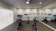 Located at Madison Square in Charlotte, NC, this single-floor fitness studio is purpose-built for cycling and group exercise. The room is thoughtfully designed with a calming aesthetic, anchored by a striking full-wall zen mural depicting smooth stones resting on tranquil water — creating an immersive, meditative atmosphere during workouts. Multiple Schwinn stationary cycling bikes are arranged across the rubber-floored studio, providing ample space for group spin sessions. A floor-to-ceiling mirror along one wall expands the sense of space and allows participants to monitor their form throughout class. The studio is equipped with a large wall-mounted display screen that can stream workout content, complemented by a smaller interactive touch-screen panel for programming and controls. A ceiling fan with integrated lighting ensures proper air circulation and comfort during high-intensity sessions. A well-organized rack of medicine balls and stability balls in varying sizes adds versatility to the workout options available. Crown molding and clean white baseboards lend a polished, upscale finish to the space, while recessed lighting throughout ensures consistent, well-distributed illumination. The room's neutral gray walls and dark rubberized flooring strike a balance between functionality and refined design, making this studio an inviting destination for fitness enthusiasts.