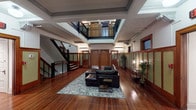 This distinctive space showcases a thoughtfully preserved interior with strong architectural character. At its heart is a grand, open common area anchored by a central skylight atrium that floods the floor with natural light from above, creating an airy, voluminous atmosphere despite the single-story layout. Rich hardwood flooring runs throughout, complementing warm-toned wood trim, wainscoting, and craftsman-style wall paneling finished in sage green and deep mahogany tones.

The main gathering area is furnished with a leather sectional sofa and a glass-top coffee table arranged atop a patterned area rug, offering a welcoming lounge setting. A striking staircase with dark metal railings and wood treads rises from the main floor, leading to an upper mezzanine level enclosed by ornate iron balustrades and overlooking the atrium below.

Along the perimeter, frosted-glass panel walls with wood framing provide subtle privacy while maintaining visual openness. Numbered solid wood doors with frosted glass sidelights line a corridor, suggesting individual suites or offices accessed from the shared common hall. Decorative corbels, transom windows, and period-appropriate sconce lighting reinforce the building's classic aesthetic throughout, lending the space a refined, professional ambiance rooted in historic craftsmanship.