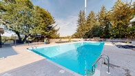 This outdoor pool facility in Fort Wayne, Indiana offers a thoughtfully designed recreational space ideal for relaxation and leisure. The centerpiece is a large, rectangular swimming pool with crystal-clear blue water, accessible via stainless steel entry rails and surrounded by a broad concrete pool deck. The deck provides ample space for circulation and lounging, with numerous modern chaise lounge chairs arranged throughout the area for sunbathing and unwinding.

A dedicated pool house anchors one end of the facility, featuring a classic white exterior with louvered ventilation panels and clearly posted pool rules and safety signage. The structure provides functional support amenities for pool users. Adjacent to the pool, a generous sandy lounge area accommodates additional rows of chaise lounges, creating a relaxed, resort-like atmosphere.

A stone accent wall lines one side of the pool deck, adding a natural, textured architectural element to the space. A grill station is also present on the deck, enhancing the facility's outdoor entertaining capabilities. The entire area is enclosed by a secure perimeter fence and is beautifully framed by tall, mature evergreen and deciduous trees that provide shade, privacy, and a lush natural backdrop. The open sky above ensures abundant natural light throughout the day.
