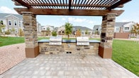 This outdoor communal space is a thoughtfully designed amenity area centered around a striking pergola structure crafted from rich, dark-stained wood beams supported by substantial natural stone columns. Beneath the pergola, a built-in outdoor kitchen anchors the space, featuring dual stainless steel grills set into a handsome stone countertop with dark work surfaces — ideal for communal gatherings and outdoor entertaining. The pergola and grilling station sit atop a generously sized paver patio composed of neutral-toned interlocking stone, providing a durable and attractive surface for outdoor seating and foot traffic. The patio transitions seamlessly into wide concrete walkways that wind through the surrounding landscape. Lush, well-maintained green lawns extend outward from the patio, framed by decorative gravel beds with newly planted trees that add a natural element to the setting. A decorative stone accent wall serves as a focal landscaping feature, adding architectural interest to the grounds. Views from the pergola look out across the community toward multi-story residential buildings with balconies, craftsman-style facades, and manicured surroundings. The open sky above and abundant natural light make this an inviting, functional outdoor destination within the community.