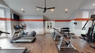 This fitness center is designed to support a comprehensive workout experience within a single, open-plan space. The room's most distinctive architectural feature is its vaulted ceiling, which rises to a generous peak and contributes to an airy, spacious atmosphere. A bold orange horizontal stripe runs along the perimeter walls, contrasting against the light gray upper walls and white beadboard wainscoting below, giving the space a clean, energetic aesthetic. Wood-look flooring runs throughout, adding warmth underfoot.

Natural light enters through multiple windows, including a curved bay window arrangement along one wall, keeping the space bright and inviting. A ceiling fan with integrated lighting provides additional air circulation and illumination, supplemented by recessed can lights. Large wall mirrors along one side visually expand the room and allow for form-checking during exercise.

The cardio zone features several treadmills equipped with digital displays, an elliptical trainer, and a row of spin bikes. A wall-mounted flat-screen television is positioned for easy viewing from the cardio equipment. The strength training area includes a multi-station cable machine suitable for a full range of resistance exercises, complemented by a tiered dumbbell rack offering multiple weight increments. French doors provide access to an adjacent entry area, and posted facility rules are clearly displayed near the entrance.