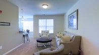 This single-story space presents an open-concept layout that seamlessly connects the living and dining areas. The living room is carpeted and comfortably arranged with a neutral-toned sofa, floral accent chairs, and a round upholstered ottoman serving as a central coffee table. A framed piece of artwork adorns the wall, adding a decorative focal point to the space. Multiple windows fitted with white horizontal blinds allow natural light to fill the room, creating a bright and welcoming atmosphere. A ceiling fan is visible toward the dining area, contributing to the overall comfort of the space. The dining area features bar-height seating and a ceiling fan, offering a casual and functional space for meals. The neutral color palette of beige and soft white tones throughout the walls and furnishings creates a cohesive, understated aesthetic. Flush-mount ceiling lighting provides consistent illumination across the living area. The overall ambiance is warm and residential, balancing practicality with comfortable, everyday living.