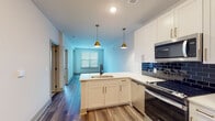 This single-floor residence offers a thoughtfully designed open-concept layout where the kitchen flows seamlessly into the living area. The kitchen serves as the focal point of the space, featuring a central peninsula with an undermount sink, crisp white shaker cabinetry accented with brushed gold hardware, and a striking deep-blue subway tile backsplash. A full suite of stainless steel appliances — including a range, over-the-range microwave, dishwasher, and refrigerator — is complemented by two pendant lights with gold-toned fixtures that add warmth to the space. Gray accent cabinetry flanking the refrigerator provides visual contrast. The open living area extends from the kitchen, bathed in natural light from large windows fitted with horizontal blinds. Durable hardwood-style plank flooring runs throughout the main living spaces, while the bedroom features plush wall-to-wall carpeting and a ceiling fan. The bathroom showcases a marble-style countertop with an undermount sink, matte black fixtures, a large framed mirror, and a tiled shower. Clean white walls and a neutral palette throughout contribute to the bright, contemporary ambiance of the space.