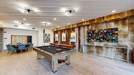 Located at 2501 N Orange Blossom Trail in Orlando, FL, this well-appointed game room serves as a vibrant communal amenity space designed for entertainment and social gathering. The single-floor layout offers a generous, open feel, with light-toned large-format tile flooring running throughout and a bright, coffered ceiling fitted with recessed lighting and track fixtures that illuminate the space evenly.

The room's most striking design feature is an accent wall composed of reclaimed wood planking and corrugated metal panels, lending an industrial-rustic character that contrasts beautifully with the clean white walls and modern finishes. Colorful, eclectic artwork is displayed throughout, adding energy and personality to the environment.

Anchoring the space is a full-size billiards table with a slate-blue felt surface, accompanied by a wall-mounted cue rack nearby. A shuffleboard table provides additional entertainment options. A cozy lounge nook along one wall features a built-in bench seat with a wall-mounted flat-screen TV above, complemented by cylindrical drum side tables. A round dining table surrounded by upholstered blue chairs offers a casual seating area, and a large wall-mounted Scrabble board adds a playful touch.

Large glass doors and windows along one side of the room flood the interior with natural light and provide direct access to an outdoor terrace area with views of palm trees and the surrounding property.