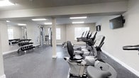Located at 2000 Birchwood Circle in Bethlehem, PA, this fitness center offers a well-equipped, single-floor workout facility designed to support a comprehensive exercise routine. The space features a clean, modern aesthetic with neutral gray walls and durable speckled rubber flooring that is both functional and easy to maintain. Recessed rectangular lighting fixtures are evenly distributed across the ceiling, ensuring bright, consistent illumination throughout the room. Natural light flows in through several windows, enhancing the open and airy atmosphere of the space. The cardio section is equipped with a row of elliptical trainers and upright exercise bikes from True Fitness, providing a solid selection of low-impact aerobic options. On the opposite side of the room, a multi-station cable machine anchors the strength training area, accompanied by a weight bench and additional free weight equipment, allowing for a full range of resistance exercises. A structural support column is integrated into the layout without disrupting the flow of the space. A wall-mounted display screen adds a modern, functional touch. Overall, the fitness center is thoughtfully arranged to maximize usable floor space while offering a diverse range of equipment suited to various fitness levels and goals.