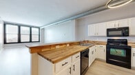 Located at 1211 West 9th Street in Cleveland's dynamic Ohio City neighborhood, this one-bedroom, one-bathroom unit blends industrial character with modern finishes. Light wood-tone luxury vinyl flooring runs throughout, complementing the neutral gray walls and crisp white trim that create a clean, contemporary aesthetic.

The open-concept kitchen is a standout feature, showcasing warm butcher block countertops that extend along a peninsula with bracket-supported bar seating — ideal for casual dining. White shaker-style cabinetry provides ample storage, while a full suite of black appliances, including a refrigerator, range, microwave, and dishwasher, adds a bold contrast. Exposed metal ductwork along the ceiling reinforces the unit's industrial loft character, and a circular pendant light adds a modern touch.

The spacious living area benefits from large black-framed windows that invite natural light and offer views of the surrounding cityscape. A well-proportioned hallway connects the main living spaces to the bedroom, which features a high clerestory window and exposed ductwork, maintaining the industrial aesthetic. Two interior doors near the bedroom provide access to additional storage or closet space.

The bathroom is efficiently appointed with a full tub-shower combo, a vanity with a light countertop, and an overhead storage cabinet, offering practical functionality in a clean, white finish.