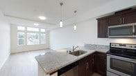This single-floor space showcases a thoughtfully designed open-concept layout that seamlessly connects the kitchen and living area. The kitchen is a standout feature, equipped with rich dark-stained cabinetry that provides a striking contrast against the light-toned granite countertops. The granite surfaces extend across a generous kitchen island, which includes a stainless steel undermount sink and a sleek pull-down faucet, along with an integrated dishwasher beneath. Two elegant cylindrical pendant lights hang above the island, complementing the flush-mount ceiling fixture that provides additional ambient lighting throughout the space. The cabinetry features brushed metal hardware, adding a contemporary touch to the overall design. The open living area beyond the kitchen benefits from large windows that invite abundant natural light and offer views of surrounding greenery. Light-toned hardwood-style flooring runs throughout, enhancing the bright and airy atmosphere. The clean white walls and minimalist architectural details contribute to a modern, welcoming ambiance suited for both everyday living and entertaining.