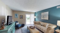 This single-floor space presents a thoughtfully designed living room that balances comfort and style. The room's most striking feature is a deep teal accent wall, which adds a bold pop of color against the warm beige tones of the surrounding walls, creating a visually dynamic yet cohesive atmosphere. A textured popcorn ceiling adds a classic touch overhead, while wall-to-wall carpeting contributes to the room's cozy, relaxed feel.

Natural light plays a central role in the space, flooding in through a large sliding glass door fitted with sheer vertical blinds. The door opens to a view of lush greenery outside, enhancing the sense of openness and providing a pleasant indoor-outdoor connection. The living area comfortably accommodates a tufted beige sofa, an upholstered ottoman serving as a coffee table, and a cushioned accent chair in navy blue, complementing the room's cool-toned palette. A turquoise sideboard and carefully selected wall art further reinforce the space's coordinated design aesthetic. A floor lamp positioned near the sliding door ensures the room remains well-lit during evening hours.