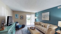 This single-floor space presents a thoughtfully designed living room that balances comfort and style. The room's most striking feature is a deep teal accent wall, which adds a bold pop of color against the warm beige tones of the surrounding walls, creating a visually dynamic yet cohesive atmosphere. A textured popcorn ceiling adds a classic touch overhead, while wall-to-wall carpeting contributes to the room's cozy, relaxed feel.

Natural light plays a central role in the space, flooding in through a large sliding glass door fitted with sheer vertical blinds. The door opens to a view of lush greenery outside, enhancing the sense of openness and providing a pleasant indoor-outdoor connection. The living area comfortably accommodates a tufted beige sofa, an upholstered ottoman serving as a coffee table, and a cushioned accent chair in navy blue, complementing the room's cool-toned palette. A turquoise sideboard and carefully selected wall art further reinforce the space's coordinated design aesthetic. A floor lamp positioned near the sliding door ensures the room remains well-lit during evening hours.