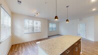 This single-floor residence showcases a thoughtfully designed open-concept layout that seamlessly connects the kitchen, dining, and living areas. The kitchen serves as a central focal point, featuring a large granite-topped island illuminated by two elegant pendant lights. Two-toned cabinetry — combining light wood upper cabinets with darker lower units — pairs with a full suite of stainless steel appliances, including a refrigerator, range, microwave, and dishwasher. Light wood-style flooring runs throughout the main living areas, contributing to a clean, contemporary aesthetic.

Multiple windows fitted with horizontal blinds flood the space with natural light, creating a bright and airy atmosphere throughout. A track lighting fixture in the living area provides additional illumination. The bedrooms feature plush carpeting and ceiling fans, with ample natural light from well-placed windows. One bedroom includes a closet and connects conveniently to the hallway.

The bathroom is finished with matching granite countertops, warm wood-tone cabinetry, a single vanity with a large mirror, and a full tub-shower combination, all set on neutral tile flooring. The overall design maintains a cohesive, modern palette of warm neutrals and contemporary finishes throughout.