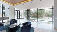This thoughtfully designed single-floor space blends contemporary architecture with refined interior details to create an inviting and functional environment. The entrance area features expansive floor-to-ceiling glass curtain walls on multiple sides, flooding the space with natural light and offering unobstructed views of the surrounding greenery and urban streetscape. Polished light-toned flooring reflects the abundant daylight throughout.

The main lounge area is anchored by plush navy blue armchairs and sofas arranged atop a rich teal area rug, creating a comfortable and visually striking gathering space. A prominent coffered ceiling panel clad in warm wood tones with integrated ambient uplighting adds architectural depth and a sense of refinement. A built-in wood shelving unit displays curated décor in coordinating blue and neutral tones, providing both visual interest and functional storage.

Adjacent to the lounge, a private dining area features a substantial wood dining table surrounded by upholstered gray chairs, illuminated by a sculptural geometric chandelier. Floor-to-ceiling glazing continues into this room, connecting occupants to the outdoor landscape. The overall ambiance is modern, welcoming, and polished.
